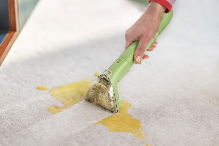 A hand holds the Little Green Mini stain tool flat against a yellow spill on carpet.