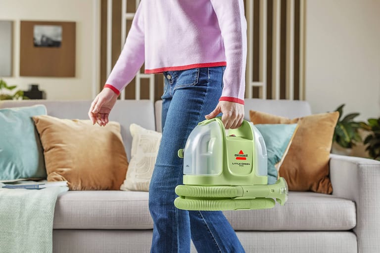 A person carries the BISSELL Little Green Mini by its top handle beside a sofa.
